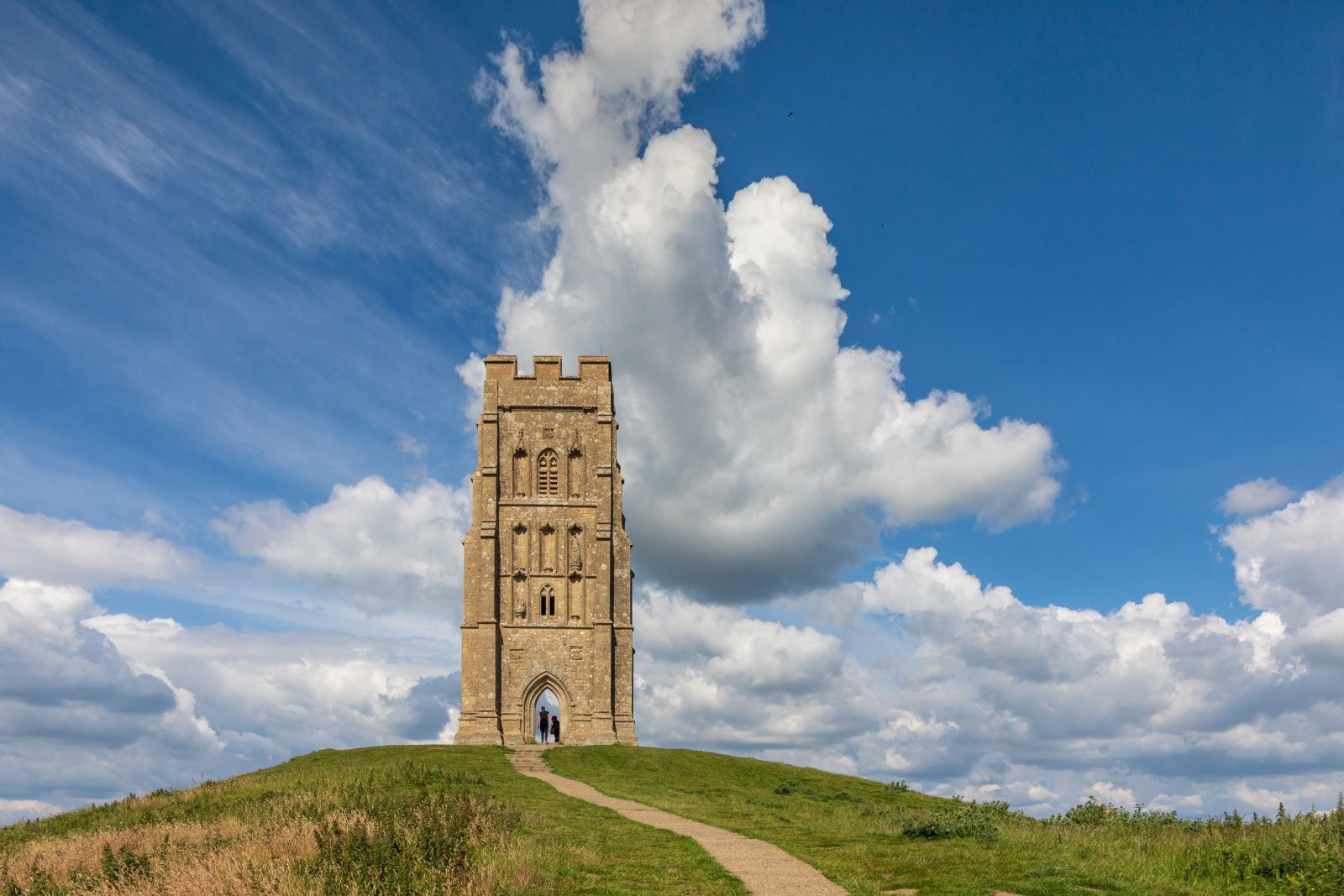 A view of a Tor with two people in the archway on top of a large gree n hill with a footpath leading to the top