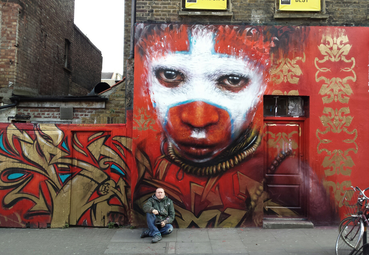 Street artist Dale Grimshaw sitting on the pavement in front of his massive and colourful street mural of a face